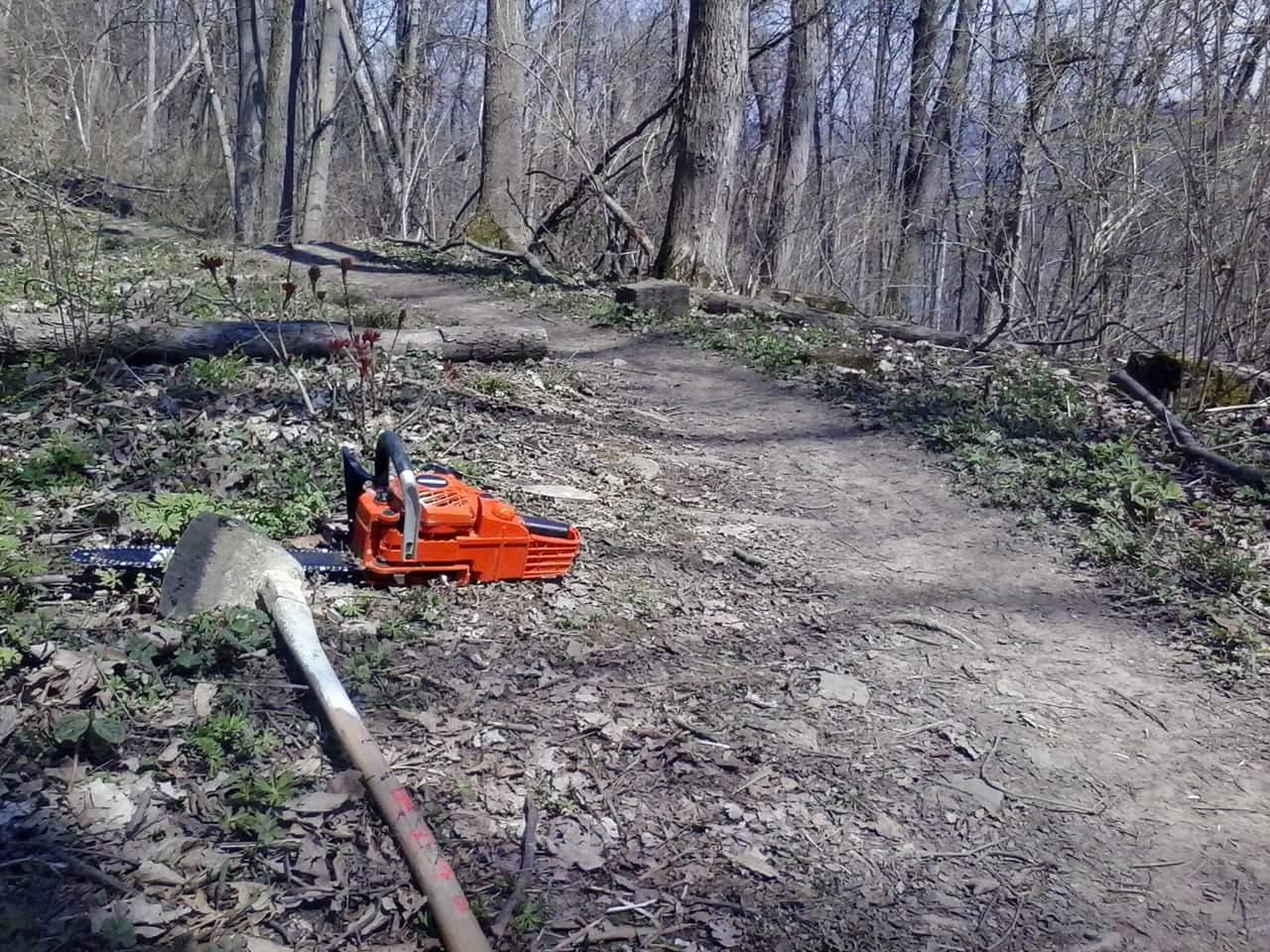 A chainsaw and a shovel resting on a dirt trail in a wooded area, surrounded by trees and sparse undergrowth. Tower Park mountain bike trail.