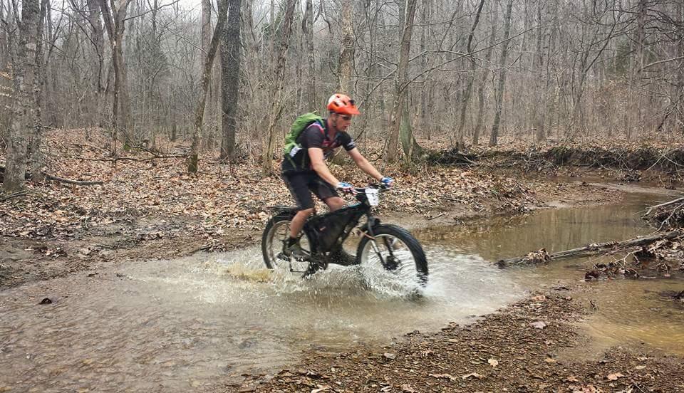 A cyclist wearing an orange helmet and a green backpack rides through a shallow stream on a muddy trail in a forested area. The scene features bare trees and fallen leaves, creating a rugged outdoor environment. Water splashes around the bike as the rider navigates the stream. Hickory Ridge mountain bike trail.