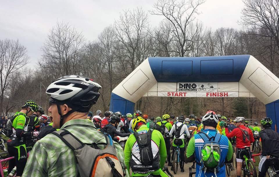 A crowd of cyclists in colorful jerseys and helmets gathered at the start and finish line of a biking event, marked by a large inflatable archway with the text "DINO." The setting features bare trees and a cloudy sky, indicating an outdoor environment, as participants prepare for the race. Hickory Ridge mountain bike trail.