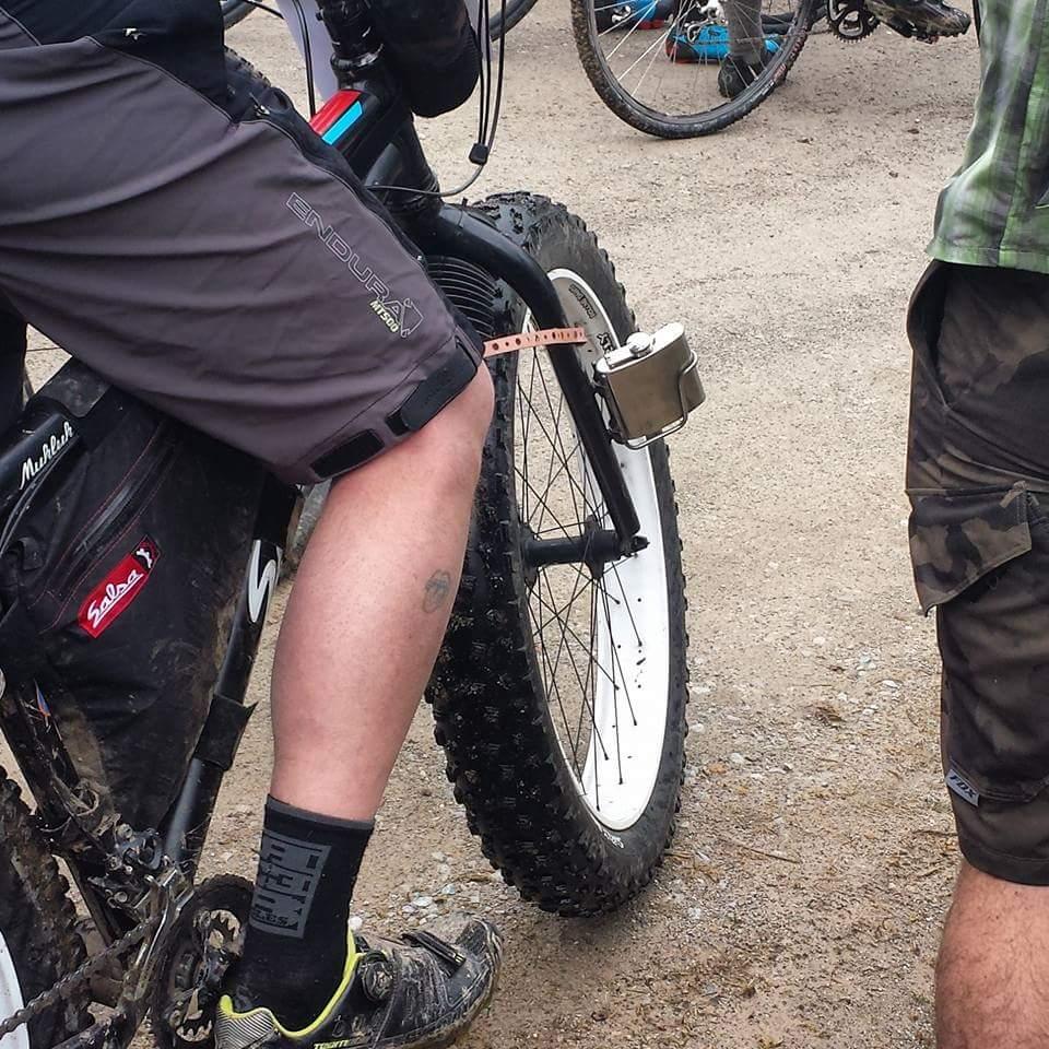 A cyclist wearing gray shorts and black socks rests on a fat-tire bike. The bike has a large white tire, and the cyclist's leg reveals a small tattoo. In the background, other bikes are visible, indicating a group or event setting. Hickory Ridge mountain bike trail.