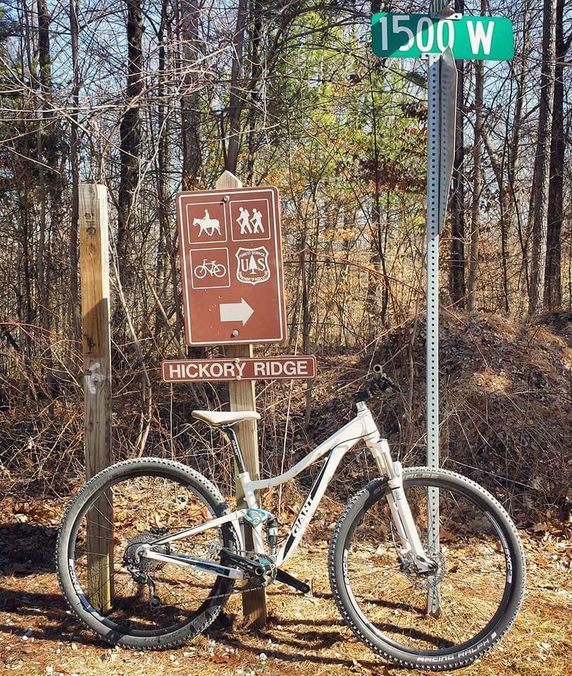 A mountain bike leans against a trail sign that indicates Hickory Ridge and features icons for horseback riding, hiking, and biking. Nearby, a street sign reads "1500 W." The background shows a wooded area with sparse foliage. Hickory Ridge mountain bike trail.