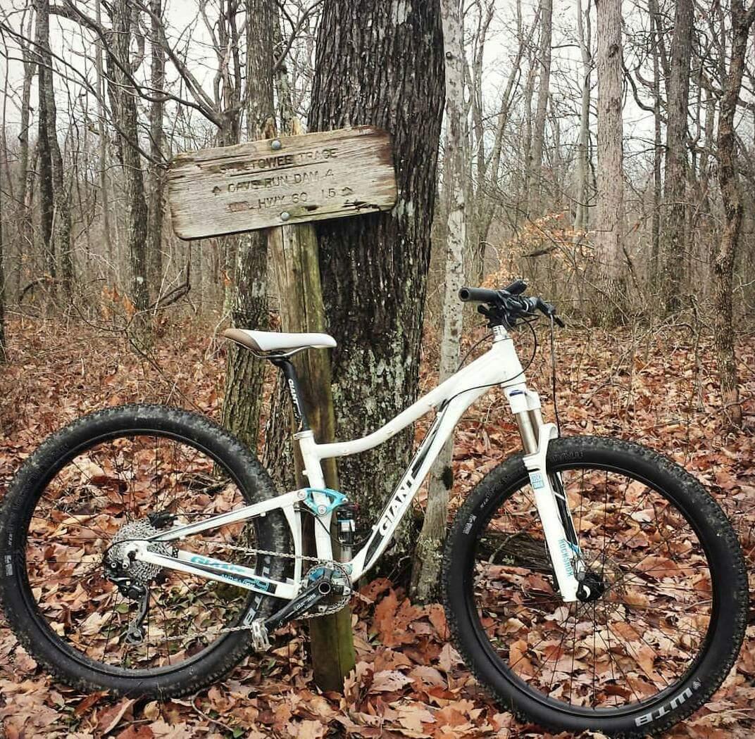 An off-road mountain bike parked beside a rustic wooden sign in a wooded area, surrounded by fallen leaves and bare trees. The sign indicates a trail and nearby highway information. Cave-run Lake mountain bike trail.