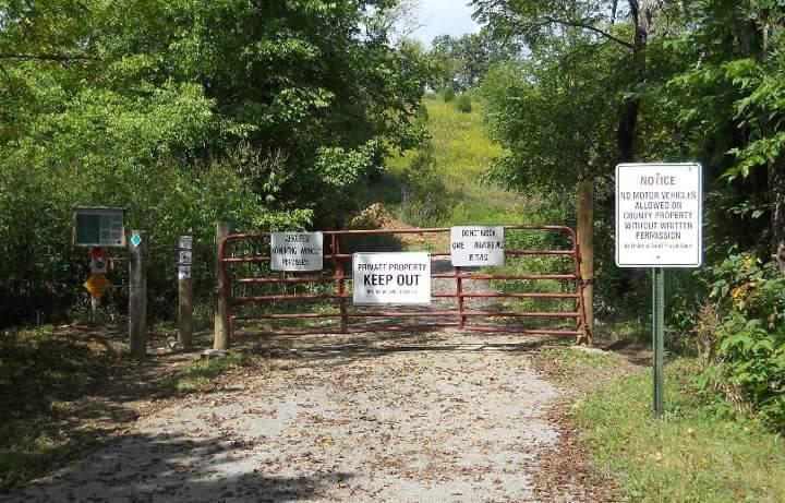 A red gate at the entrance of a private property, surrounded by lush green trees. Signs on the gate indicate "PRIVATE PROPERTY KEEP OUT" and additional notices regarding access restrictions for motor vehicles and permission requirements. The ground is covered with leaves, suggesting a wooded area leading further into the property. Skullbuster mountain bike trail.