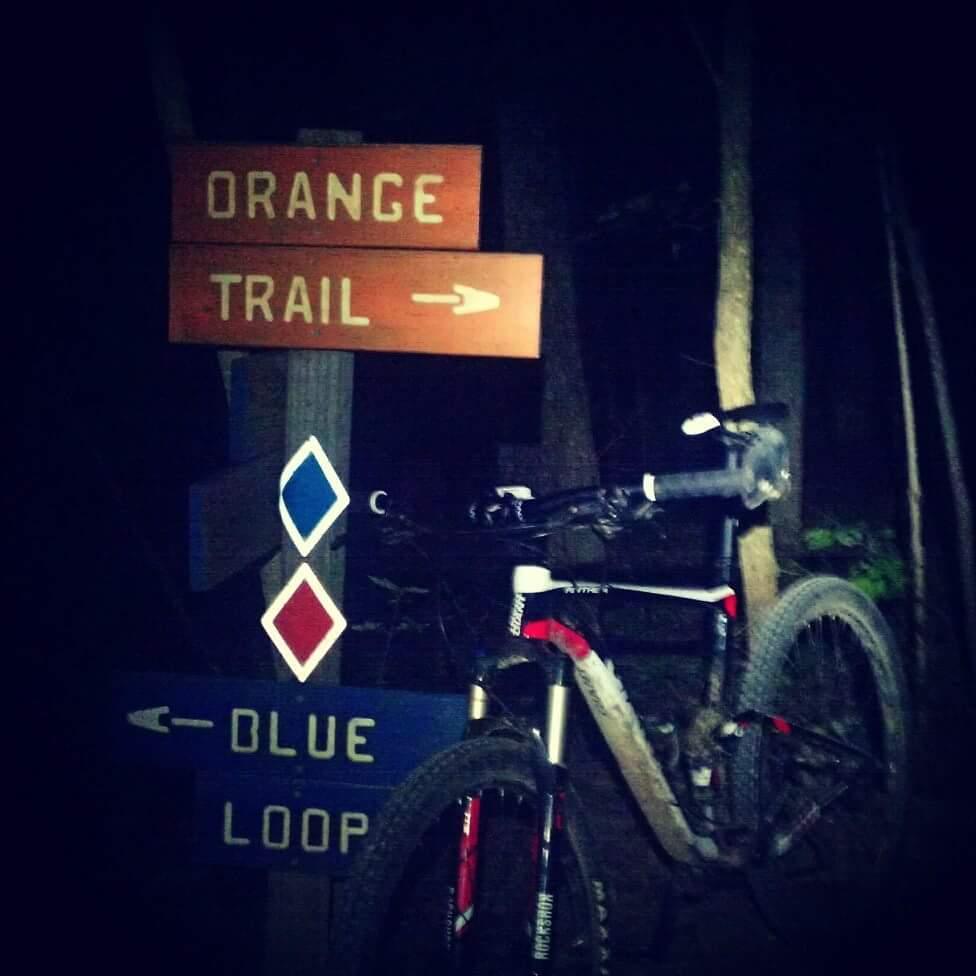 A mountain bike parked next to trail signs indicating the direction for the "Orange Trail" and "Blue Loop," set in a dark, wooded area. Skullbuster mountain bike trail.