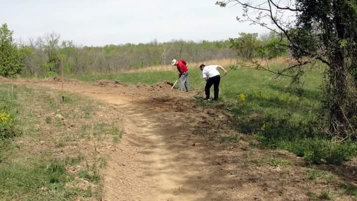 Two people working together to landscape a dirt path in a grassy area, surrounded by trees and open fields under a clear sky. One person is wearing a red shirt and the other a white shirt, both using tools to shape the path. England Idlewild Mountain Biking Park mountain bike trail.