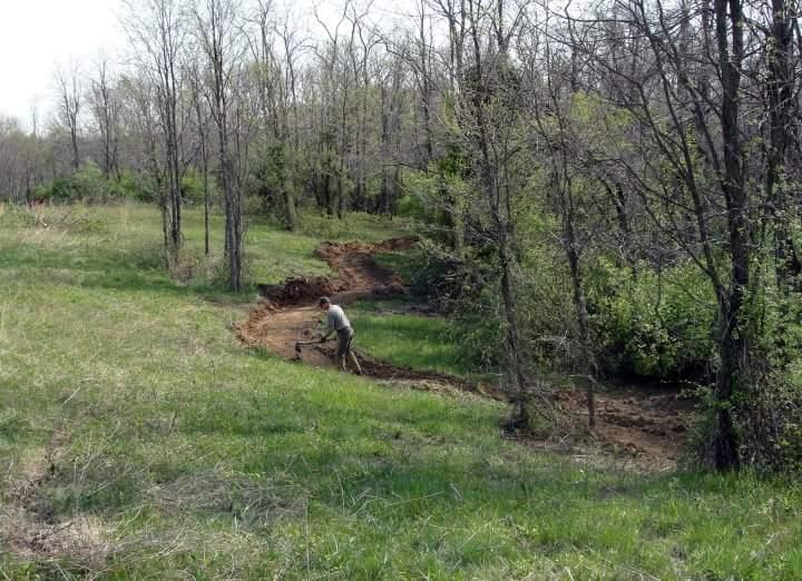 A person digging in a grassy area surrounded by trees. The landscape features uneven terrain with a path being cleared in the soil, indicating outdoor land maintenance or gardening activities. England Idlewild Mountain Biking Park mountain bike trail.