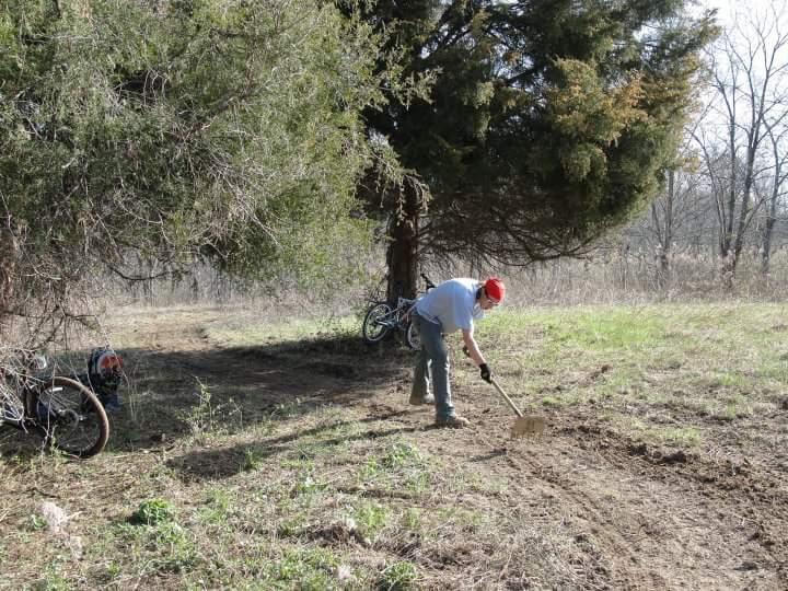 A person wearing a red hat and gloves is digging in the ground with a shovel under a large tree. In the background, there are two bicycles leaning against the tree, and the setting is a grassy area with sparse trees in the distance, indicating a rural outdoor environment. England Idlewild Mountain Biking Park mountain bike trail.