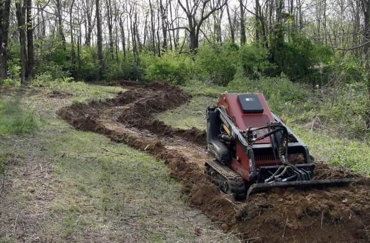 A compact track loader working on a dirt path in a wooded area, creating a winding trail through freshly disturbed soil and greenery. England Idlewild Mountain Biking Park mountain bike trail.
