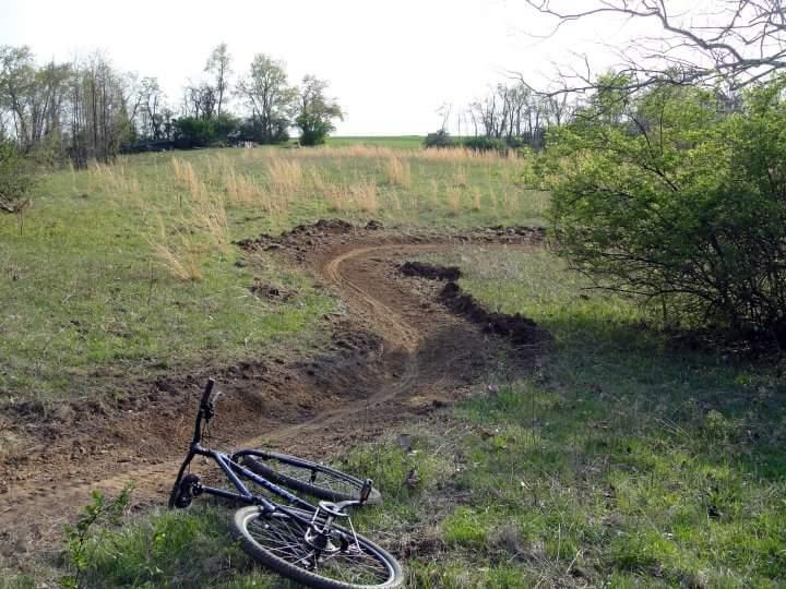 A mountain bike lies on its side near a dirt path in a grassy field, featuring a curved trail surrounded by sparse vegetation and trees in the background under a clear sky. England Idlewild Mountain Biking Park mountain bike trail.