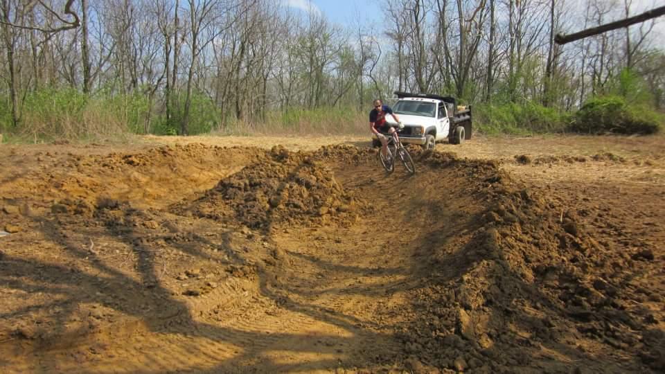 A cyclist rides a mountain bike on a dirt track that has been recently shaped, with mounds of earth nearby. In the background, a white pickup truck is parked on the side of a cleared area with sparse trees. The scene is set in a natural outdoor environment on a sunny day. England Idlewild Mountain Biking Park mountain bike trail.