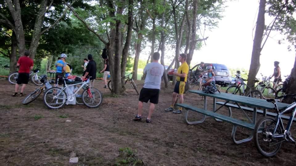 A group of cyclists gathered in a wooded area, with several bicycles parked nearby. Some individuals are interacting with each other, while others are seated at a picnic table. The scene captures a casual outdoor gathering, surrounded by trees and natural greenery. England Idlewild Mountain Biking Park mountain bike trail.
