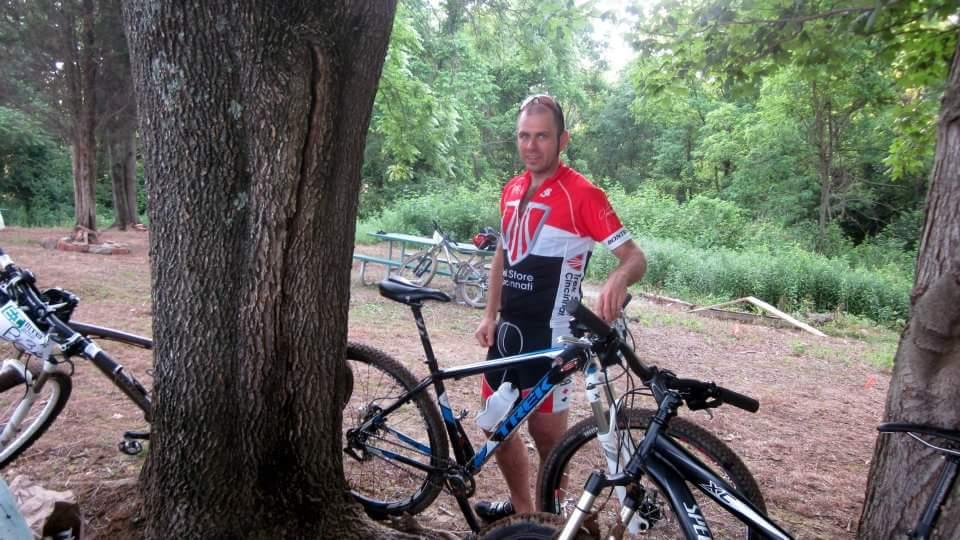 A man in a red and white cycling jersey stands next to his mountain bike, leaning against a tree in a wooded outdoor setting. Several other bikes are visible in the background, surrounded by greenery. England Idlewild Mountain Biking Park mountain bike trail.