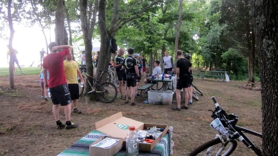 A gathering of cyclists in a wooded area, with some people standing around a picnic table. Bicycles are parked nearby, and a variety of refreshments are laid out on the table. The scene is lively, with individuals wearing cycling gear and casual clothing, enjoying the day in a green, natural setting. England Idlewild Mountain Biking Park mountain bike trail.