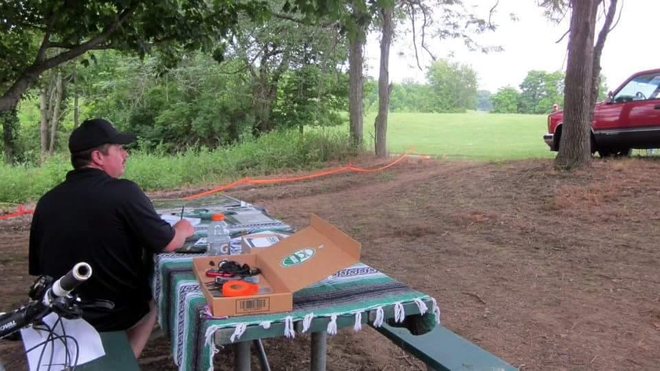 A person sitting at a picnic table under a tree, looking out towards a grassy field. The table is covered with a patterned cloth and has various items, including a pizza box and some tools. A bicycle is leaning against the table, and a red pickup truck is parked nearby. The scene is set in a natural, wooded area. England Idlewild Mountain Biking Park mountain bike trail.