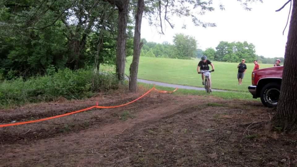 A mountain biker navigates a dirt trail marked with bright orange tape, surrounded by trees. In the background, several spectators watch from near a parked red truck, with a grassy field and a path visible in the distance. England Idlewild Mountain Biking Park mountain bike trail.