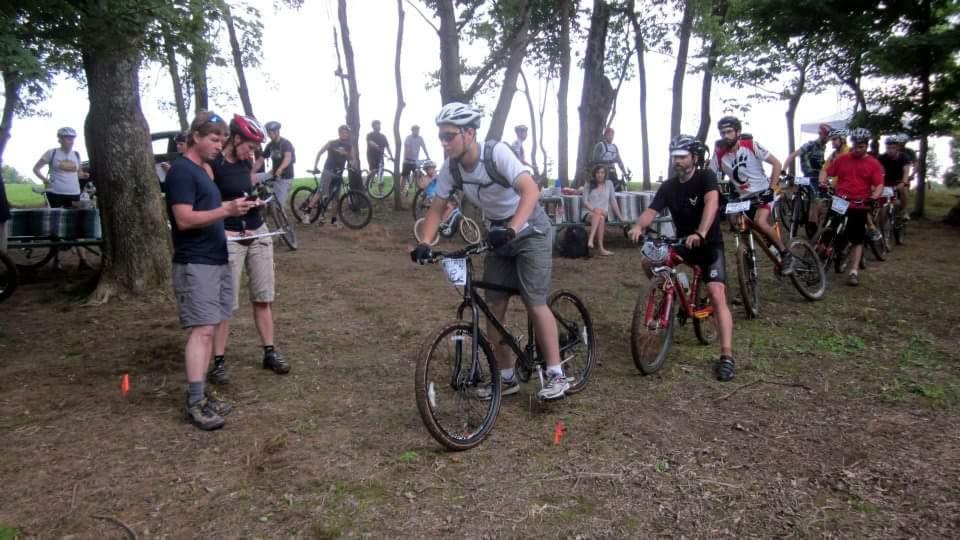 A group of mountain bikers prepares for a race in a wooded area. Some participants are mounted on their bikes, while others are chatting and checking equipment. A few people can be seen in the background, and the atmosphere is lively and focused, typical of a pre-race setting. England Idlewild Mountain Biking Park mountain bike trail.