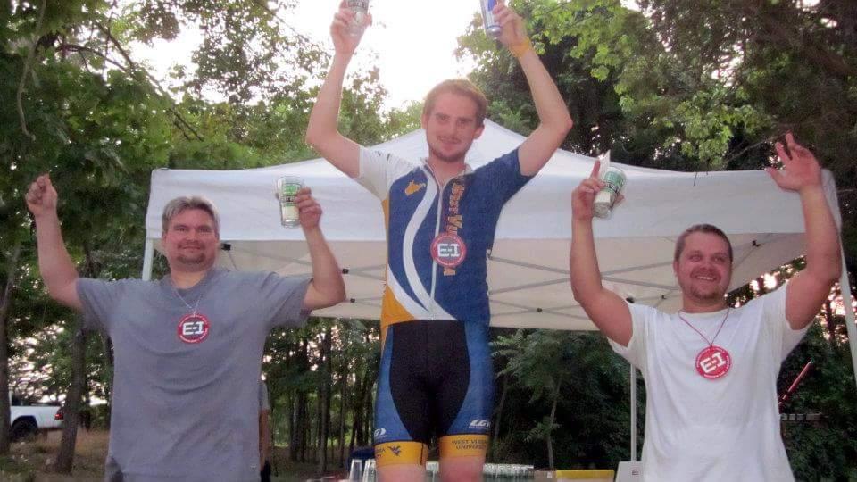 Three men celebrate on a podium, raising their arms cheerfully while holding drinks. The center figure, wearing a colorful cycling jersey and shorts, stands above the others, who are in casual t-shirts. All three have medals around their necks and appear to be celebrating a victory. A white tent and trees are visible in the background. England Idlewild Mountain Biking Park mountain bike trail.