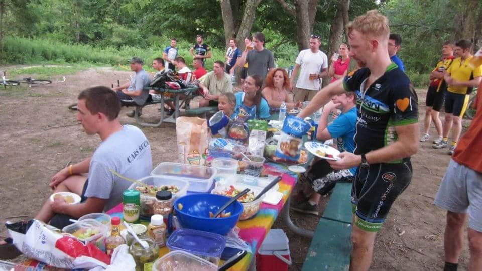 A group of cyclists and spectators enjoy a picnic in a wooded area. In the foreground, a man in cycling gear serves himself food from a colorful table filled with various dishes and snacks, while another man sits nearby with a plate of food. In the background, several people relax and chat, some sitting on benches and others standing, amidst bike gear and nature. England Idlewild Mountain Biking Park mountain bike trail.