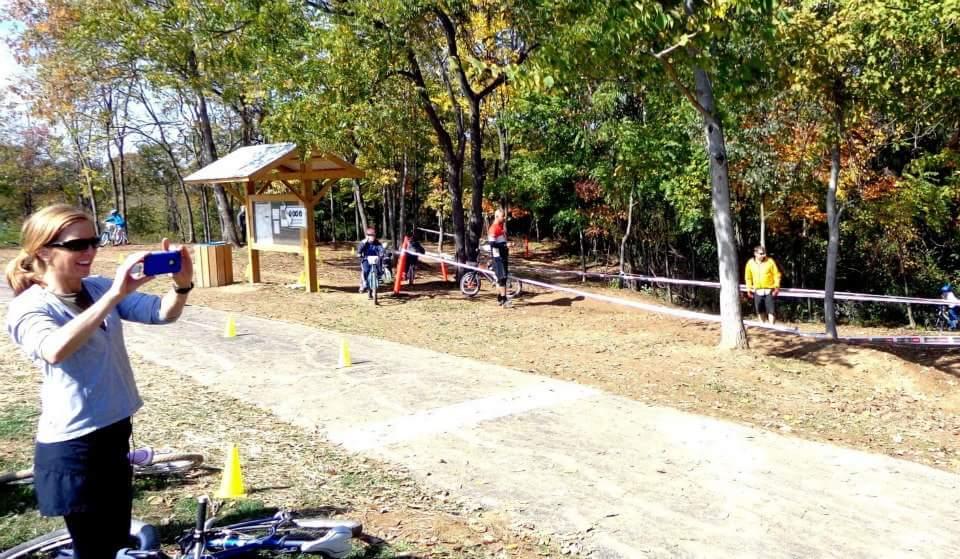 A woman taking a photo with a smartphone while standing next to a bike path. In the background, other individuals are riding bicycles and walking along a tree-lined area with autumn foliage. There are traffic cones set up along the path, and a covered structure that appears to contain informational signage. England Idlewild Mountain Biking Park mountain bike trail.