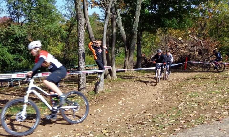 A group of cyclists riding on a dirt path surrounded by trees. One cyclist, wearing a red and black jersey, is in the foreground, while others follow behind. A child is seen playfully perched on a tree branch in the background, with a pile of branches nearby. The scene showcases a sunny day with scattered autumn leaves on the ground. England Idlewild Mountain Biking Park mountain bike trail.