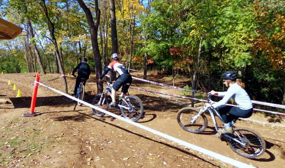 Three mountain bikers maneuver along a dirt path in a forested area during a fall day. They are surrounded by colorful autumn foliage, and the track is marked with cones and ropes. The cyclist in front wears a black outfit, while the second wears a jersey with a red and white design. The third cyclist is a woman in a light blue long-sleeve shirt, all wearing helmets for safety. England Idlewild Mountain Biking Park mountain bike trail.