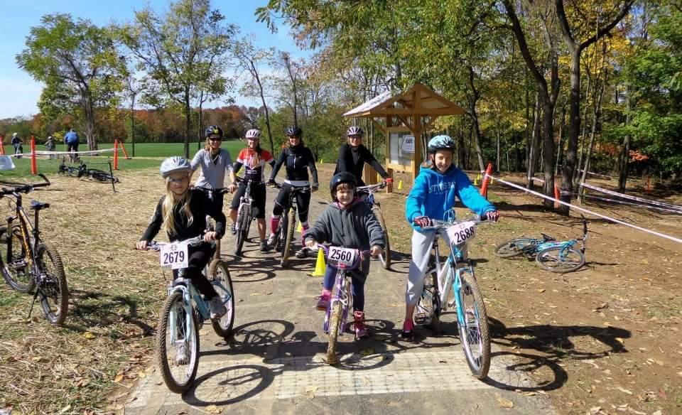 A group of young cyclists, including two girls in the foreground, are lined up on a dirt biking trail in a wooded area, ready for a race. They are wearing helmets and numbered race bibs, and several bicycles are positioned nearby. In the background, other participants can be seen, along with trees showing autumn foliage. A wooden structure indicating a checkpoint is also visible. England Idlewild Mountain Biking Park mountain bike trail.