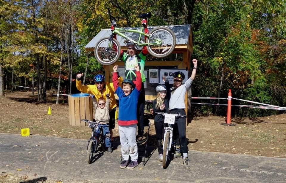 A group of six enthusiastic cyclists, celebrating their achievements in a forested area. One cyclist is holding a green bicycle above their head, while others cheer and pose with their own bikes. The scene is vibrant with fall foliage in the background, and includes a wooden structure displaying race information. England Idlewild Mountain Biking Park mountain bike trail.