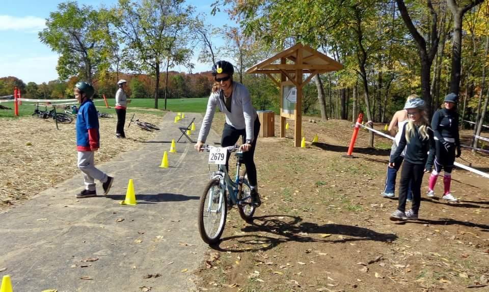 A participant rides a bicycle on a path marked with cones at an outdoor cycling event. Surrounding them are spectators, including children and adults, with several bicycles parked nearby. The scene is set in a park with trees and colorful autumn foliage in the background, capturing a sunny day filled with activity. England Idlewild Mountain Biking Park mountain bike trail.