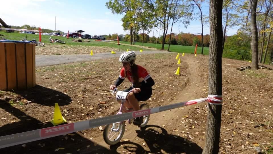 A young child in a helmet and cycling gear rides a balance bike along a dirt path, navigating a course marked with yellow cones. The background features trees with autumn foliage and a scenic grassy area, while a barrier tape with the word "Trek" is visible in the foreground. England Idlewild Mountain Biking Park mountain bike trail.