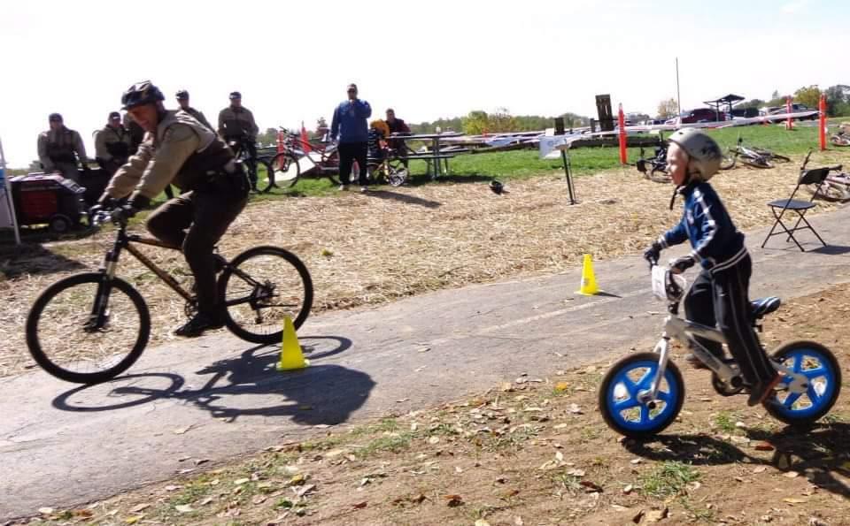 A police officer riding a bicycle navigates a course while a young child on a balance bike follows closely behind. The scene takes place outdoors, with spectators in the background and a grassy area visible. Traffic cones are set up along the path, indicating a biking event or practice session. England Idlewild Mountain Biking Park mountain bike trail.