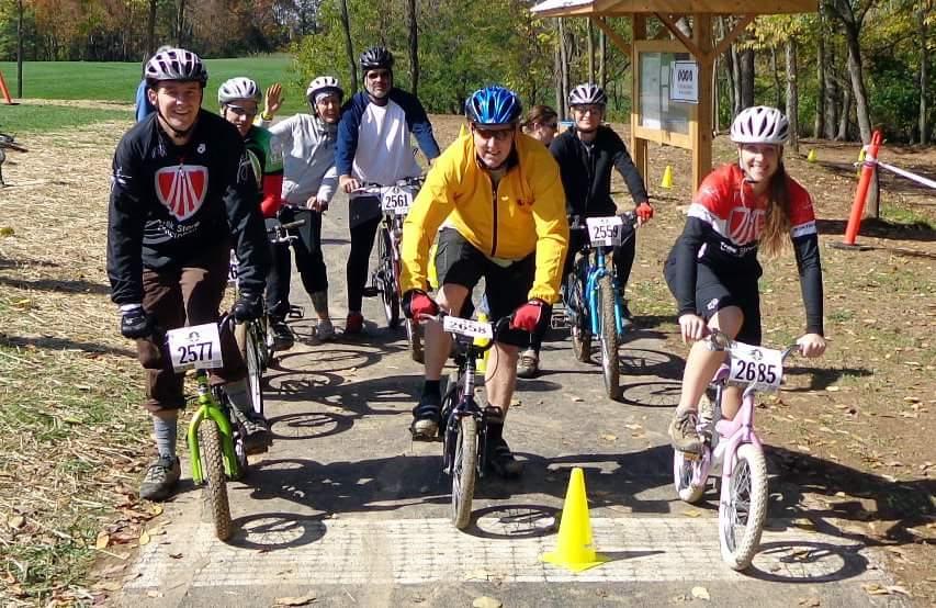 A group of cyclists, including adults and children, is gathered at the starting line of a biking event. They are wearing helmets and numbered bibs, with several bicycles of various sizes and colors. A yellow cone marks the beginning of the course, and a wooden structure in the background appears to serve as a registration or timing station. The setting features trees and a grassy area, indicating an outdoor environment during a sunny day. England Idlewild Mountain Biking Park mountain bike trail.