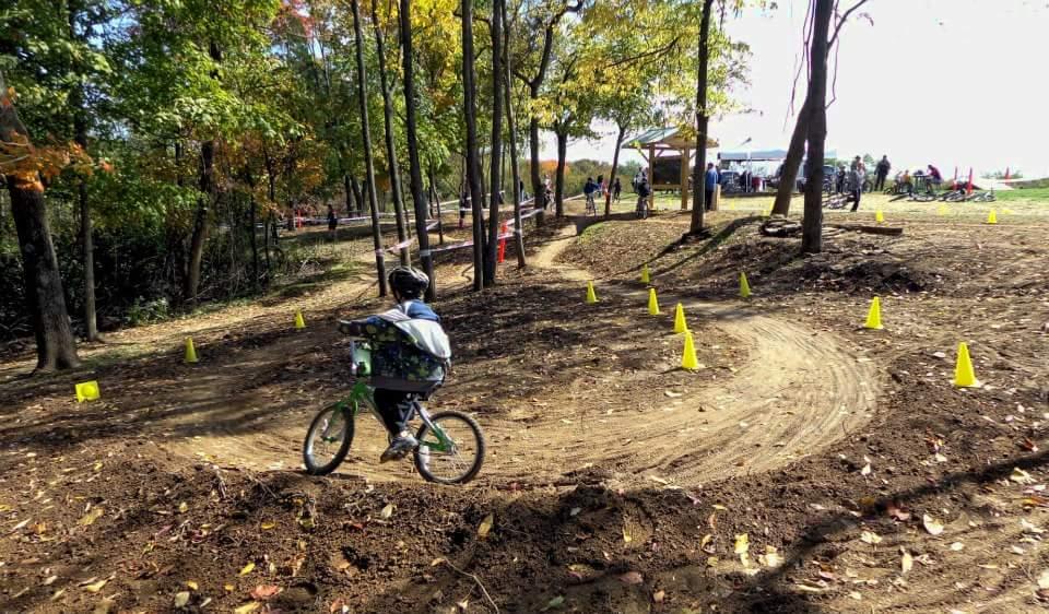 A young child riding a green bicycle on a dirt path surrounded by trees, with yellow cones marking the course. The scene captures a fall day, featuring colorful autumn leaves and a small gathering of people in the background. England Idlewild Mountain Biking Park mountain bike trail.