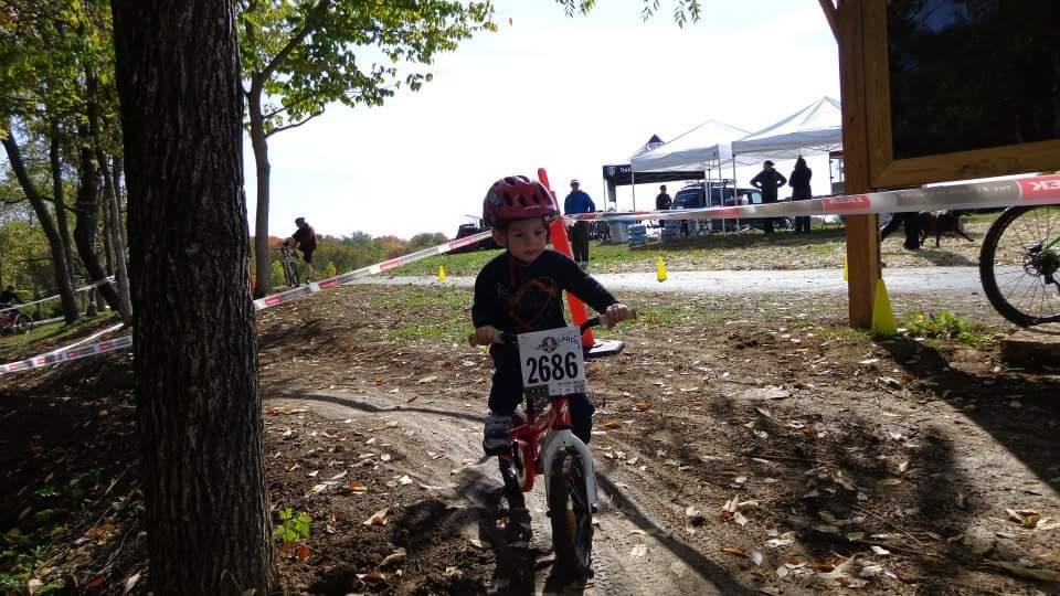 A young child wearing a red helmet rides a small bicycle on a dirt path, surrounded by trees and colorful foliage. The child has a race number displayed on their bike. In the background, there are tents and people observing the event, with another cyclist in motion nearby. The scene captures a sunny day and the excitement of a bike race. England Idlewild Mountain Biking Park mountain bike trail.