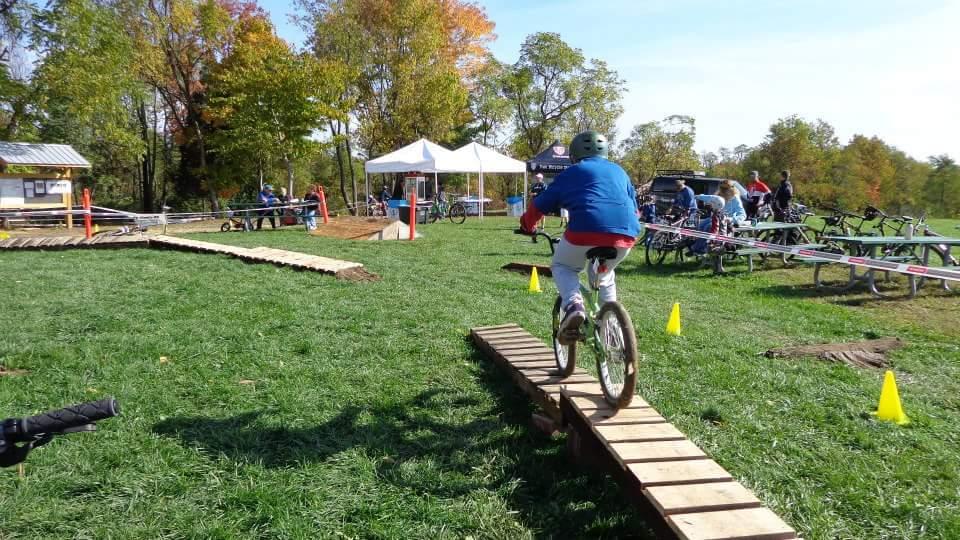 A person riding a mountain bike navigates a wooden plank bridge in a grassy outdoor area. In the background, there are tents, bicycles, and other cyclists, with colorful trees indicating autumn foliage. England Idlewild Mountain Biking Park mountain bike trail.
