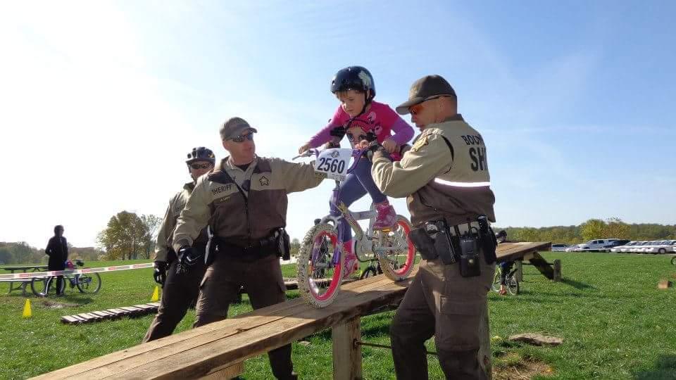 A young girl wearing a pink helmet and a pink shirt balances on a bicycle as she rides across a narrow wooden beam. Two uniformed officers assist her, helping to ensure her safety during the activity. In the background, other children and bicycles can be seen in a grassy area under a clear blue sky. England Idlewild Mountain Biking Park mountain bike trail.
