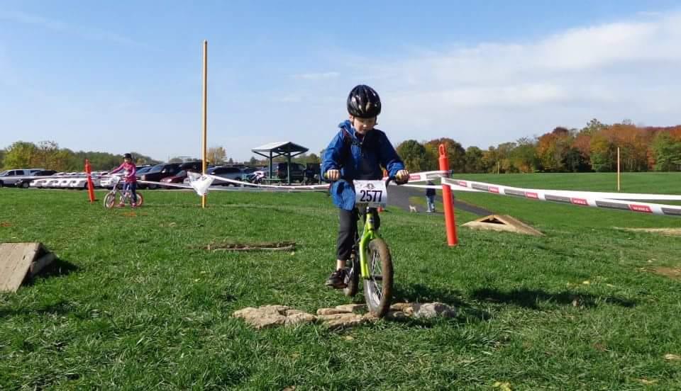 A young child in a blue jacket and black helmet rides a green bicycle over a small rocky obstacle in a grassy area. In the background, another child on a bicycle is visible, along with parked cars and a shelter under a clear blue sky. The scene captures a moment from a cycling event. England Idlewild Mountain Biking Park mountain bike trail.