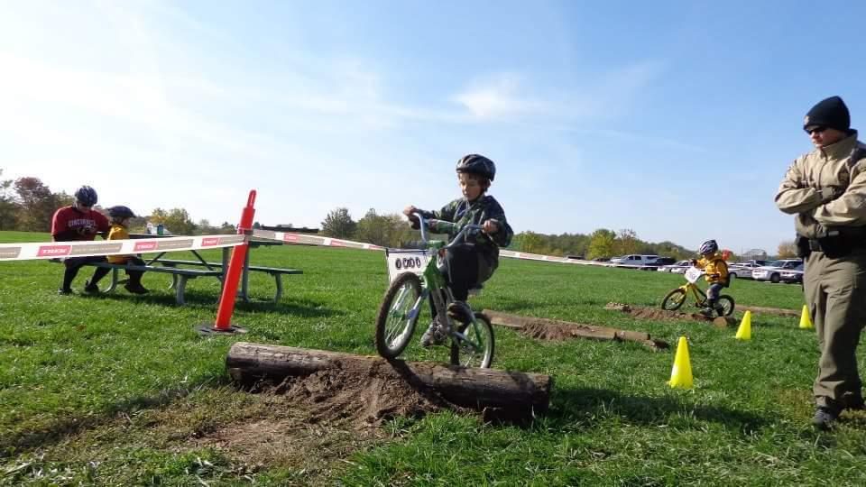 A young child riding a green bicycle navigates over a log on a grassy outdoor course, while a man watches nearby. In the background, other children are sitting by a picnic table and another child is riding a bike, with yellow cones marking the course. The scene takes place on a bright, sunny day. England Idlewild Mountain Biking Park mountain bike trail.