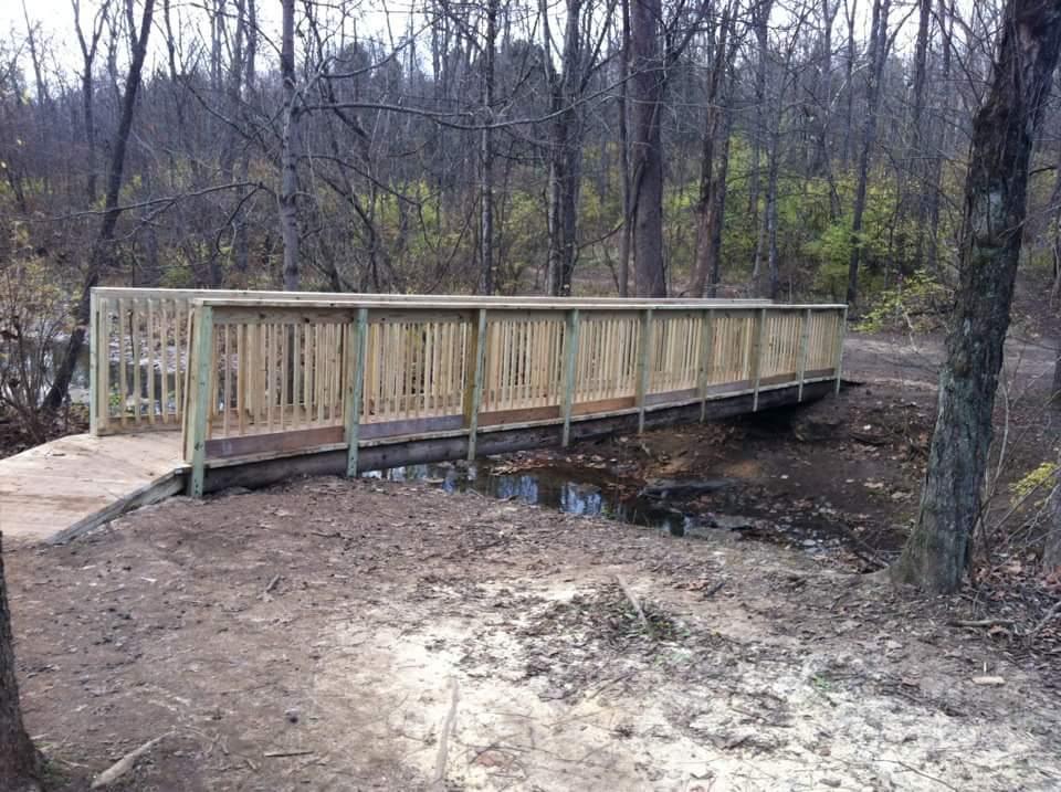 A wooden bridge with railings, spanning a small stream in a forested area. The surroundings include bare trees and some greenery, indicating early spring or late fall. The ground is a mix of dirt and grass. England Idlewild Mountain Biking Park mountain bike trail.