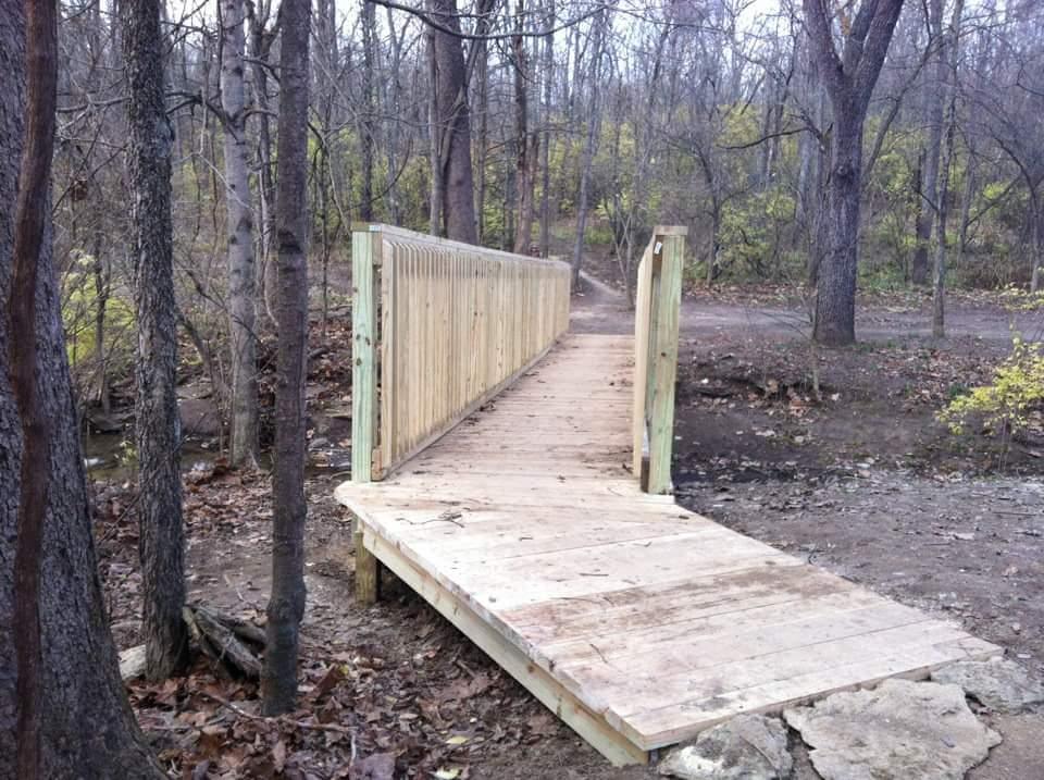 A wooden footbridge leading through a wooded area, surrounded by trees and fallen leaves. The bridge has a simple railing on either side and is positioned over a dry, dirt path. The scene is serene and evokes a sense of nature and tranquility. England Idlewild Mountain Biking Park mountain bike trail.