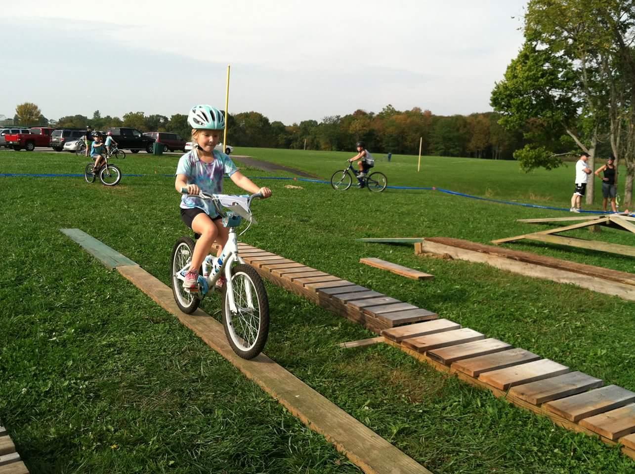 A young girl rides a bicycle across a wooden plank path set up on grass, wearing a helmet and a colorful shirt. In the background, other children are also biking, while adults watch nearby. The scene is outdoors, with trees and a clear sky visible in the distance. England Idlewild Mountain Biking Park mountain bike trail.