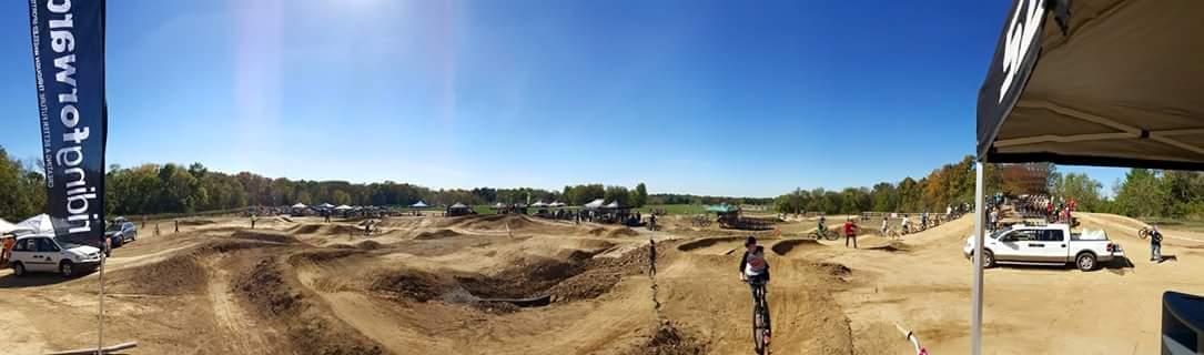 A panoramic view of a BMX track on a clear day, featuring multiple dirt jumps and trails. In the foreground, a cyclist is riding along the track, while other riders and spectators are visible in the background near tents and parked vehicles. The scene is set against a bright blue sky with minimal clouds. England Idlewild Mountain Biking Park mountain bike trail.