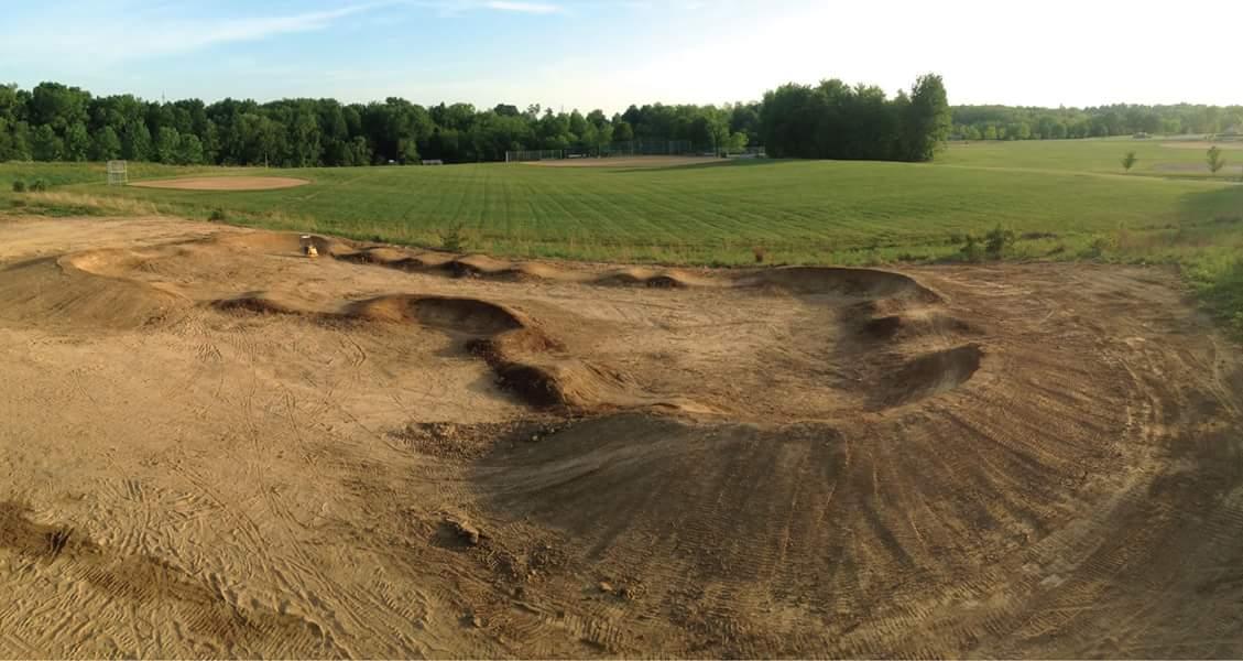 A dirt bike track featuring a series of mounds and dips, situated in an open field surrounded by trees. In the background, a well-maintained grassy area and a baseball diamond are visible, under a clear sky. England Idlewild Mountain Biking Park mountain bike trail.