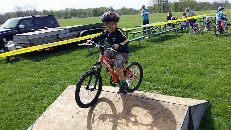 A young child wearing a helmet rides a bicycle over a small ramp in a grassy area, with spectators in the background and picnic tables lined up nearby. England Idlewild Mountain Biking Park mountain bike trail.