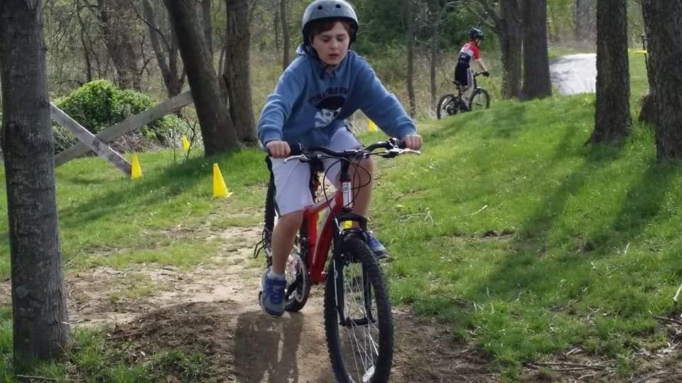 A child wearing a helmet rides a red bicycle along a dirt path in a green park, focused on navigating the trail. In the background, another child on a bike can be seen. Yellow cones are placed along the path, indicating an area for riding. Trees line the path, and the scene captures a sunny day in the outdoors. England Idlewild Mountain Biking Park mountain bike trail.