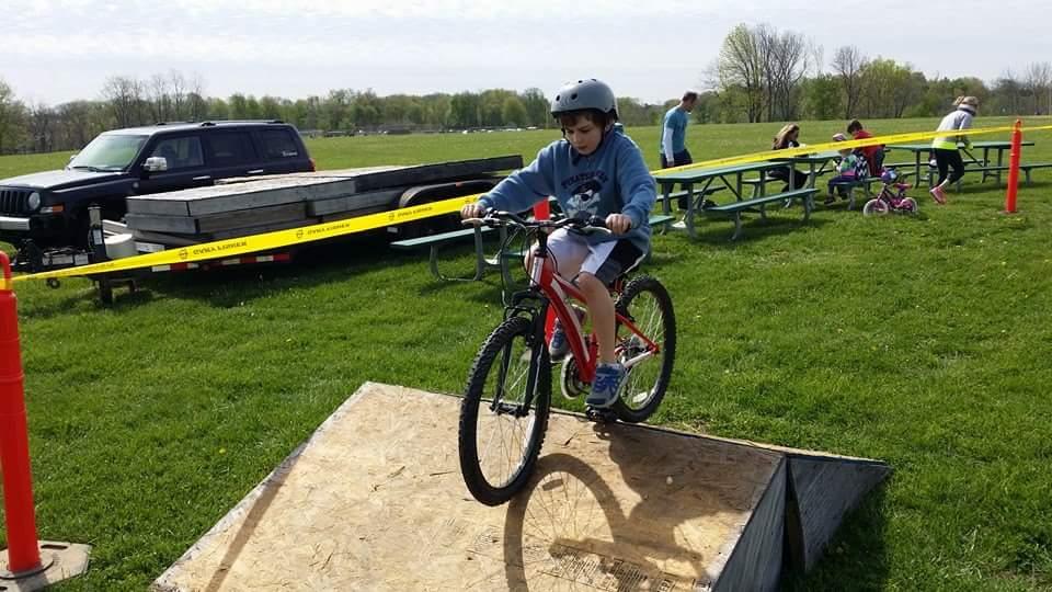 A young boy wearing a helmet rides a red bicycle as he approaches a wooden ramp in a grassy outdoor area. Several people and parked vehicles are visible in the background, along with picnic tables. The scene captures a lively atmosphere, indicating a day of outdoor activities. England Idlewild Mountain Biking Park mountain bike trail.