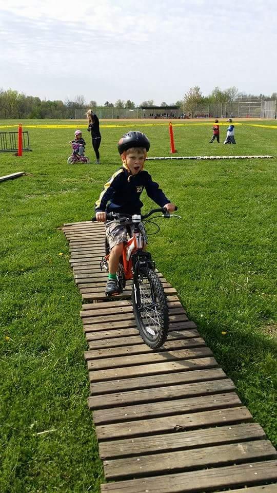 A young child riding a bicycle on a wooden plank path in a grassy park. The child is wearing a helmet and has an enthusiastic expression. In the background, other children can be seen riding bikes and playing, with trees and a sports field visible under a partly cloudy sky. England Idlewild Mountain Biking Park mountain bike trail.