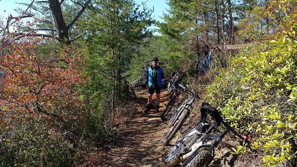 A cyclist stands on a narrow dirt path surrounded by trees and colorful shrubs. Several mountain bikes are propped against the greenery, indicating a biking adventure. The scene is set on a sunny day, enhancing the vibrant colors of the foliage. Big South Fork mountain bike trail.