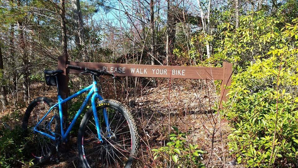 A blue bicycle parked next to a wooden sign that reads "PLEASE WALK YOUR BIKE," surrounded by a wooded area with green shrubbery and trees. Big South Fork mountain bike trail.