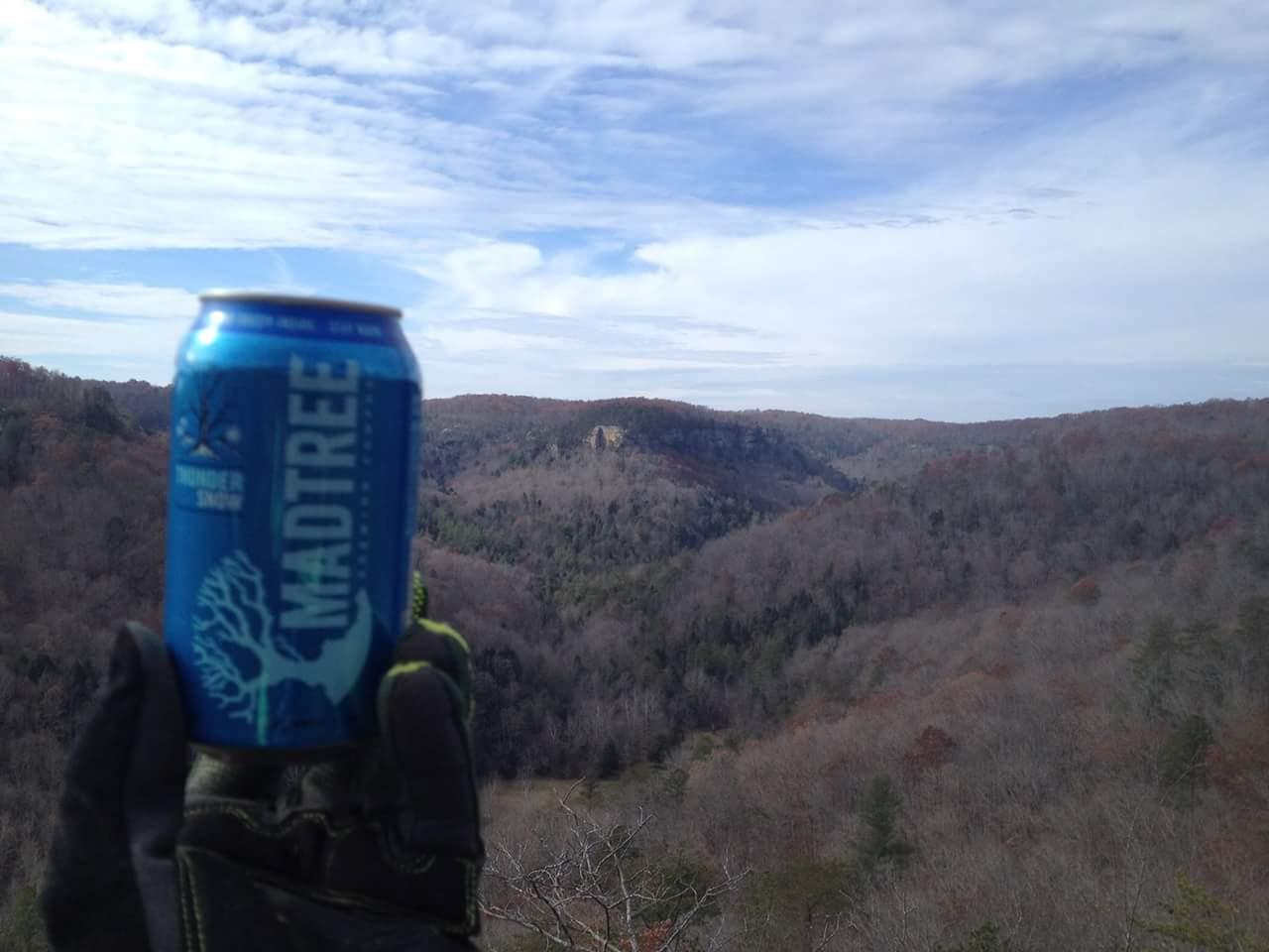 A person's gloved hand holding a can of MadTree beer in front of a scenic view of rolling hills and a partly cloudy sky. The landscape features trees in varying shades of brown, indicating autumn or early winter. Big South Fork mountain bike trail.