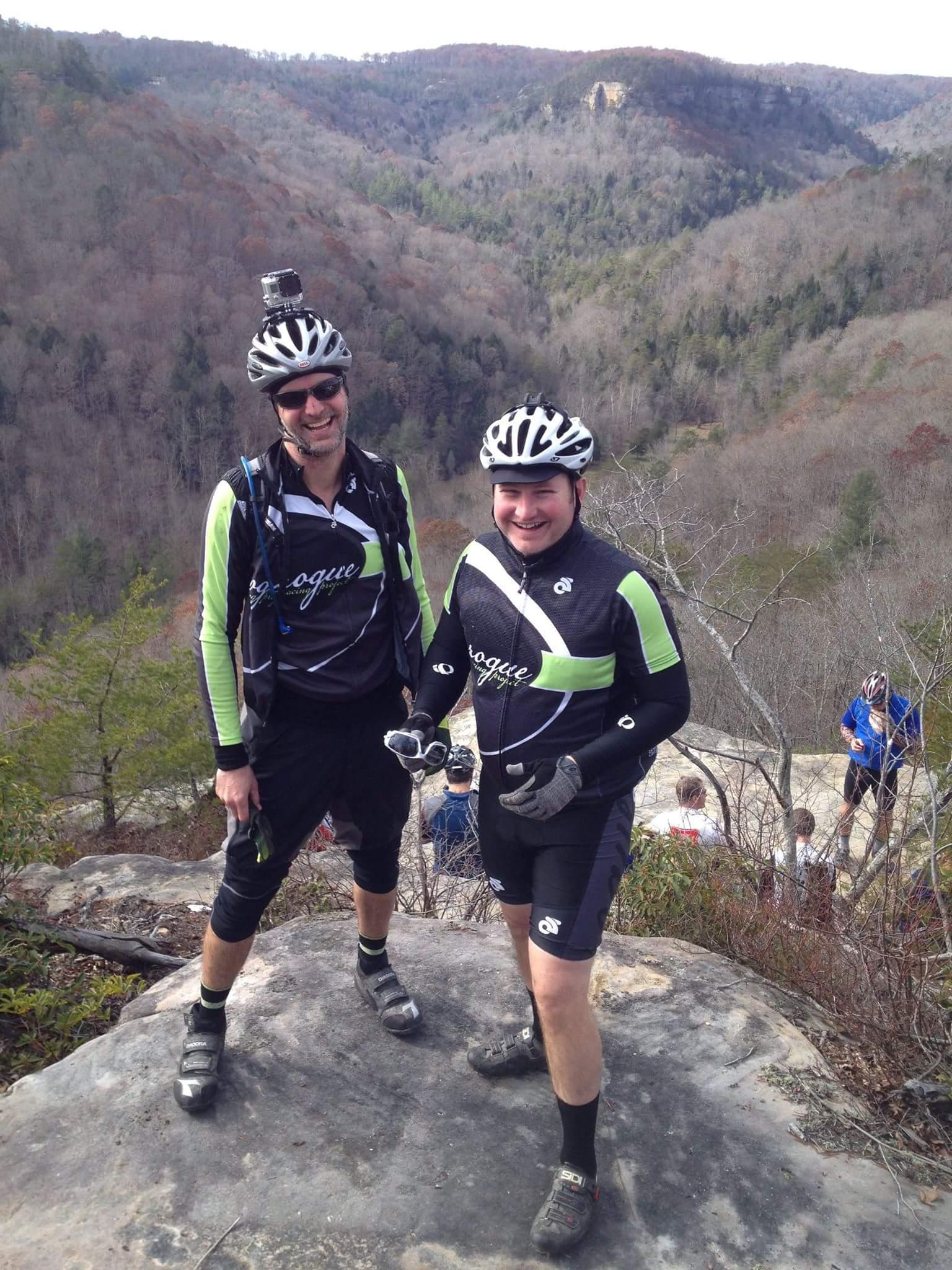 Two cyclists in bright, matching outfits stand on a rocky outcrop with a scenic view of rolling hills in the background. One cyclist is wearing sunglasses and has a camera mounted on their helmet, while both are posing happily for the photo. Other bikers can be seen in the distance among the trees. Big South Fork mountain bike trail.